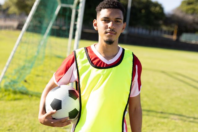 This image depicts a confident soccer player holding a soccer ball while standing on an outdoor field. He wears a training bib, indicating he is in the middle of a practice session. Ideal for use in sports advertisements, soccer training promotions, or articles about team sports and athletic confidence.