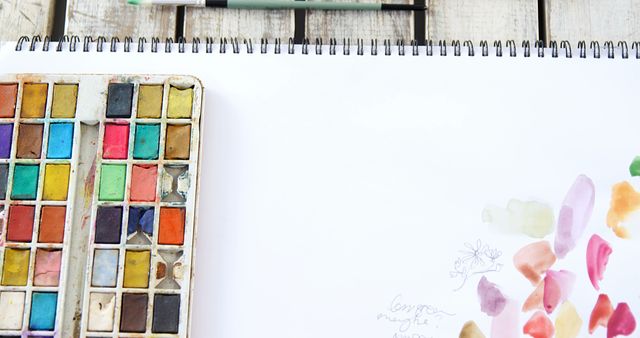 Visible watercolor palette with assorted colors beside an open sketchbook on a rustic wooden table. Ideal for projects relating to art creation, artist studios, and illustrating artistic environments.