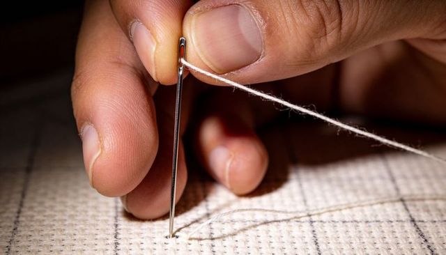Male hand threading metal needle through gridded Aida fabric for cross stitch closeup
