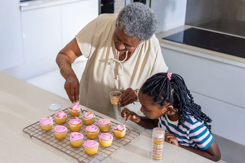 Grandmother and granddaughter decorating pink frosted cupcakes together in modern kitchen
