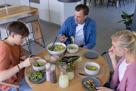 Family sharing pesto pasta at wooden dining table in modern kitchen, casual home meal