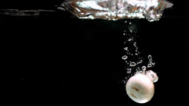 A mushroom is captured falling through water in slow motion against a sleek black background. The image showcases the dynamic motion with air bubbles forming beautifully around the mushroom. This visual is ideal for culinary blogs, artistic food galleries, health and nutrition articles, as well as promoting freshness in produce advertisements.