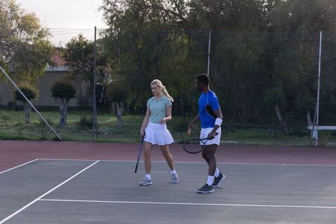 Mixed-gender tennis teammates walking on hard court at golden hour holding rackets