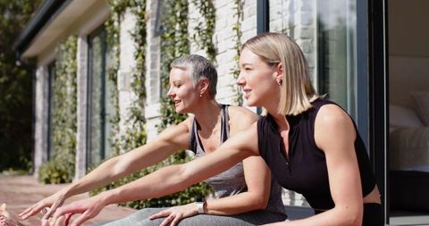 Two women stretching on sunlit patio in athletic wear with smartwatch