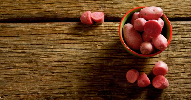 This image depicts an overhead view of a bowl filled with red skinned potatoes placed on a rustic wooden table. Some whole and cut red potatoes lie scattered around the bowl. Ideal for use in articles or websites related to cooking, healthy eating, organic farming, or vegetable farming, as well as for recipe blogs, food markets, and harvest festivals.