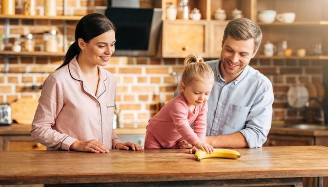 Young parents watching toddler reaching for banana on rustic kitchen island