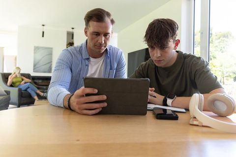 Father guiding teenage son using tablet for studying in modern sunlit home, tutoring
