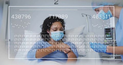 Masked patient receiving bedside care while nurse monitoring vitals in hospital ward
