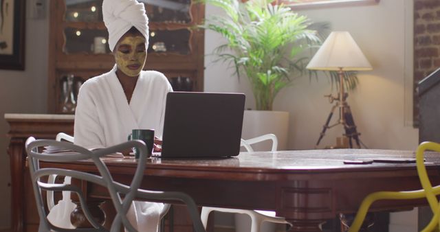 African american attractive woman with applied face mask drinking coffee and using laptop at home. beauty, pampering, home spa and wellbeing concept.