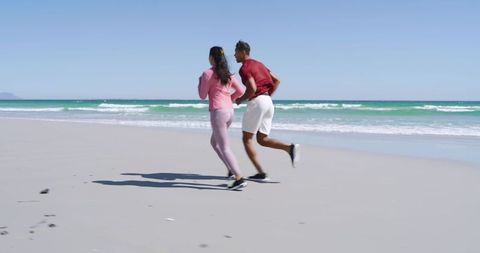 Diverse couple jogging on sunlit sandy beach by turquoise waves for outdoor fitness training