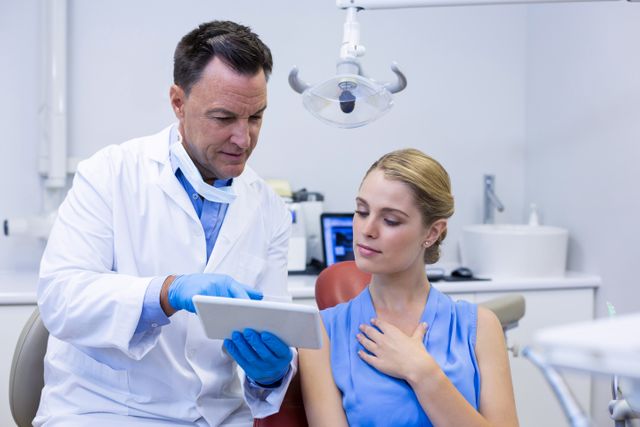 Dentist in white coat and blue gloves showing treatment plan on digital tablet to female patient in dental clinic. Ideal for use in healthcare, dental care, technology in medicine, patient consultation, and professional medical services contexts.