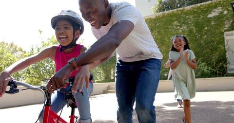 Father Teaching Daughter to Ride Bike While Sibling Cheers