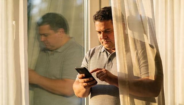 Middle-aged man checking smartphone by sunlit window with reflection and sheer curtains