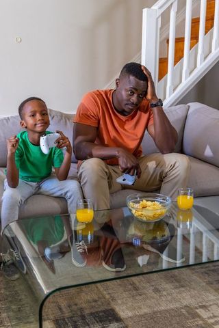 African american father and son playing video games on sofa sharing snacks, orange juice