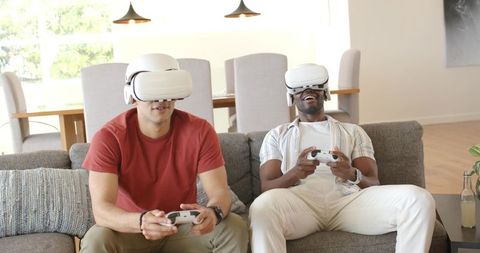 Friends playing VR games on gray sofa wearing white headsets and holding controllers smiling