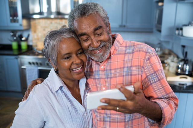 Senior couple standing in kitchen, smiling while taking selfie. Perfect for use in advertisements, articles, or blogs about senior lifestyle, family bonding, technology use among elderly, and home life.