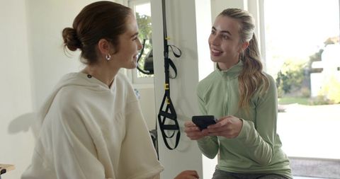 Female friends chatting in athleisure by home gym trx holding smartphone