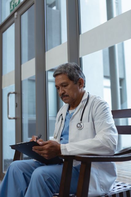 Side view of mature male doctor writing on clipboard while sitting on chair in lobby at hospital