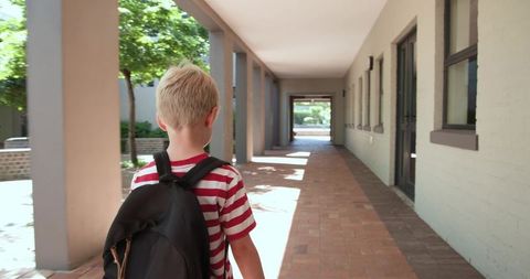 Blond schoolboy walking along campus colonnade with backpack and red striped shirt