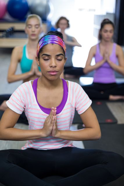 Group of women engaged in a yoga class focusing on meditation and balance. The bright gym setting and athletic clothing suggest an active, health-oriented lifestyle. Ideal for illustrating concepts of group fitness, well-being, and mindfulness in fitness marketing, wellness blogs, and meditation apps.