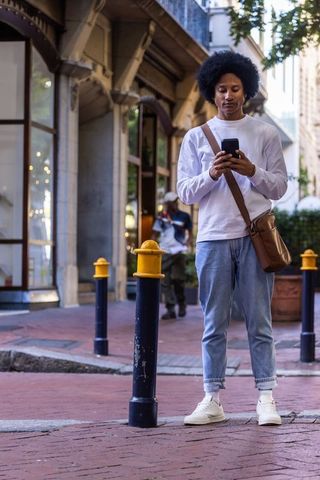 African american man checking smartphone on urban brick sidewalk with crossbody bag
