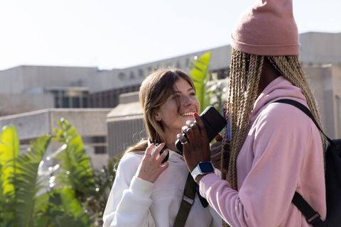 Multiracial college friends sharing music on smartphone, laughing outdoors on campus plaza