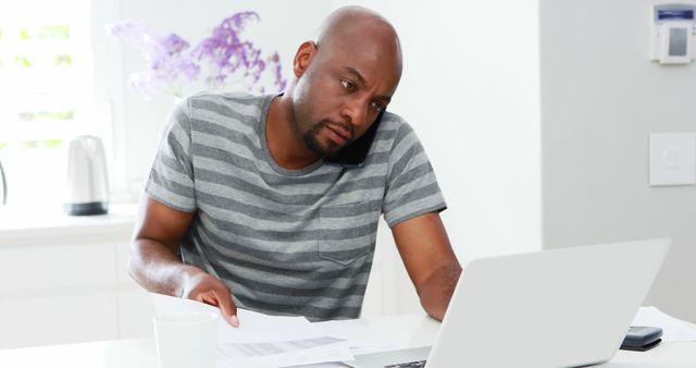 Man multitasking by talking on phone while working on laptop, suitable for articles on remote work trends, home office productivity, balancing work and communication, or modern work-from-home lifestyles.