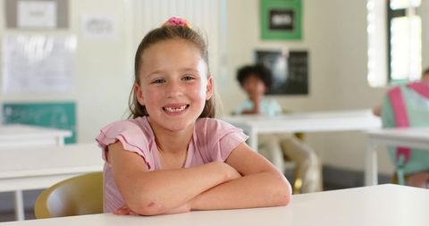 Smiling preteen girl at front desk in modern classroom wearing pink top and scrunchie