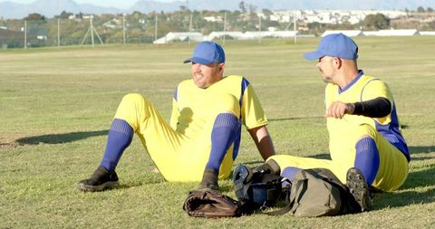 Sunlit baseball teammates sitting on grass wearing yellow uniforms and blue caps