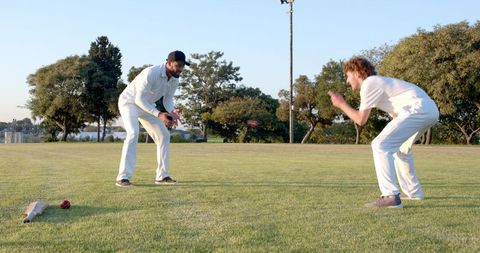 Two cricketers practicing catching and fielding drill on sunlit grass wearing white kit