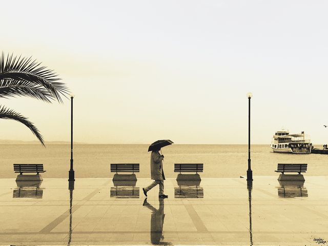 Person walking alone with umbrella on beach promenade during rainy weather. Reflections on wet floor enhance sense of solitude and reflection. Suitable for themes of loneliness, contemplation, rain, coastal life, and calming scenes.