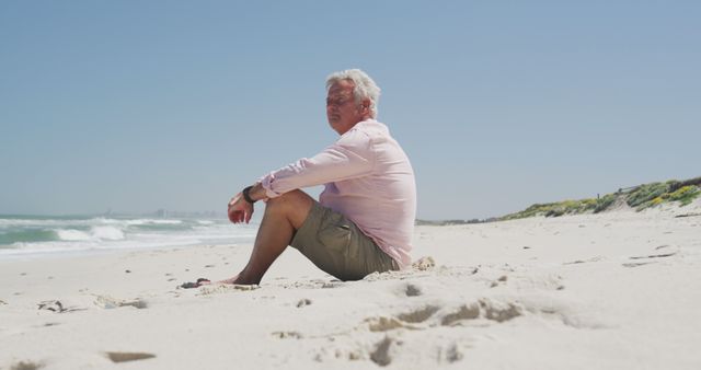 Senior man sitting alone on sandy beach looking at horizon. Ideal for use in projects depicting relaxation, retirement, peacefulness, solitude, and leisure time.