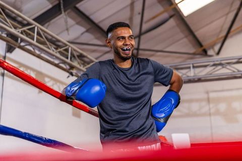 Confident male boxer leaning on ropes in gym wearing blue gloves and mouthguard