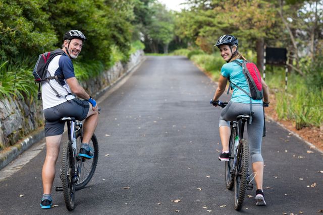 Athletic couple enjoying a bike ride on an open road, both smiling and wearing helmets. Ideal for promoting outdoor activities, fitness, healthy lifestyles, and recreational sports. Perfect for use in advertisements, blogs, and social media posts related to cycling, adventure, and active living.