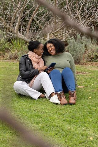African american women sharing smartphone and laughing while sitting on grass in park