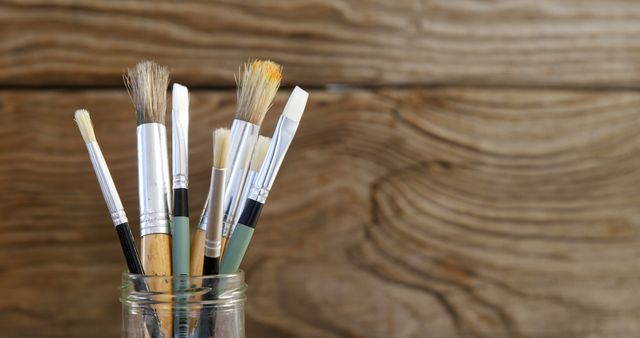 This image shows paintbrushes of various sizes and shapes arranged in a glass jar against a wooden background. Ideal for use in art and craft blogs, educational content about painting, art supply store advertisements, and any content related to creativity and craftsmanship.