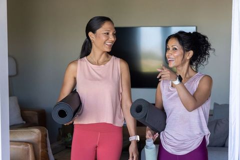 Two diverse women carrying yoga mats and water bottles smiling and chatting at home