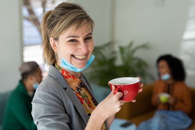 A caucasian woman at the office with her facemask pulled down holding a cup of coffee smiling. behind her are her colleagues sitting on the couch talking during their coffee break.