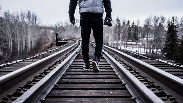 Lone traveler walking on railway tracks, holding a camera, in an outdoor setting surrounded by bare trees under an overcast sky. Ideal for themes of exploration, solitude, travel, adventure, and photography. Can be used in travel blogs, adventure magazines, or promotional material for outdoor gear.