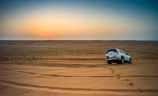 SUV driving across a vast desert at sunset with visible vehicle tracks in the sand. Ideal for illustrating concepts related to adventure travel, explorations, and off-road expeditions in remote areas. Perfect for travel blogs, adventure magazines, automotive advertisements focusing on durability and rugged terrain, and promotional material for outdoor activities.