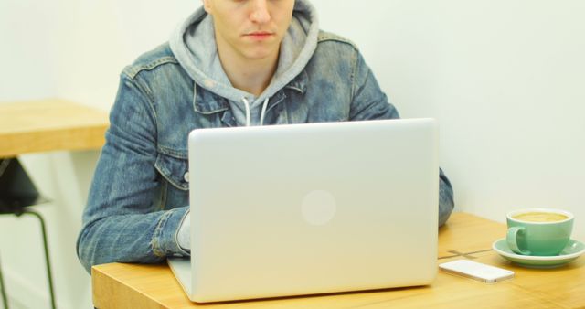 Young man is working on a laptop while seated at minimalist cafe table. Table holds a coffee cup and mobile phone. Useful for concepts like remote work, studying, casual work environment, freelancer lifestyle, technology usage, or student life.