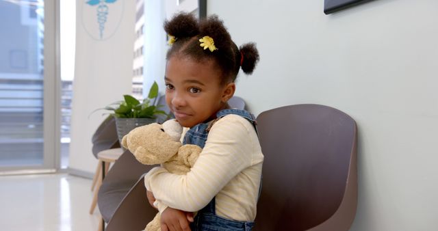 Young girl with pigtails sits in medical waiting room clutching a teddy bear, wearing denim overalls and yellow shirt. She appears a little anxious but calm. This visual is effective for content related to pediatric healthcare, hospital environments, child patients, and waiting experiences in clinics.