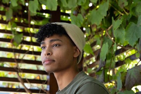 Thoughtful young african american man wearing beige beanie looking left in green pergola