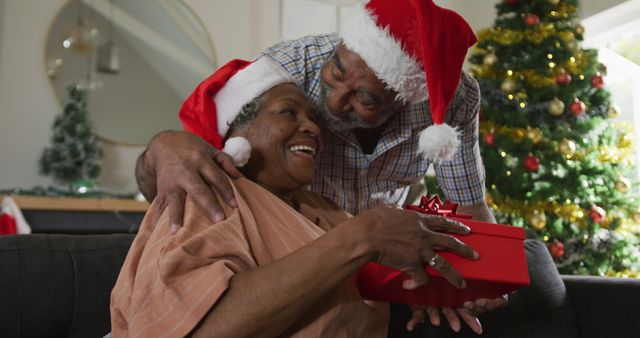 Elderly couple enjoying a joyful Christmas exchange with Santa hats, a wrapped present, and decorated tree in background. Perfect for promoting holiday family gatherings, elder care facilities, and festive cards.