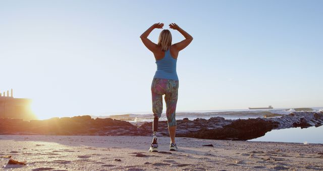 Woman with prosthetic leg wearing sportswear holding hands above head while exercising on serene beach at sunrise. Perfect for themes on resilience, fitness, inspirational stories, health and wellness campaigns, and promoting an active lifestyle.