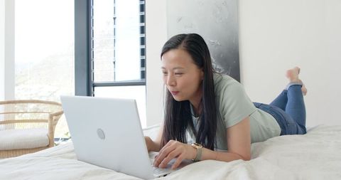 Asian woman working on laptop in modern sunlit bedroom remote work lifestyle minimalist