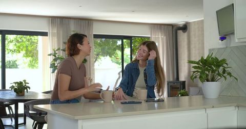 Sunlit modern kitchen friends chatting over coffee at island, holding mugs and phones