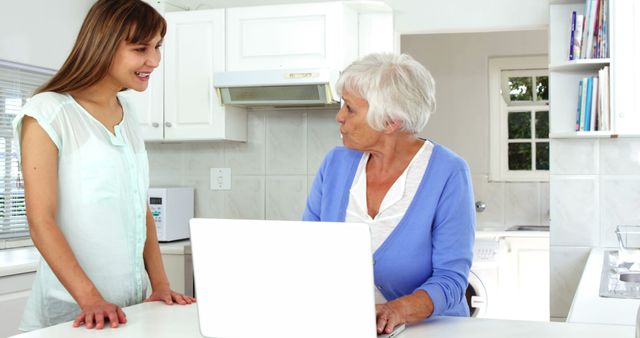 This image shows a young woman helping an elderly woman use a laptop in a bright, modern kitchen. Suitable for depicting family bonding, technology use among seniors, or intergenerational learning.