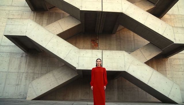 Solitary woman in red gown standing before brutalist concrete zigzag staircase, minimalist symmetry