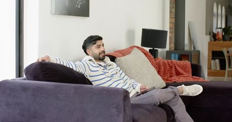 Young indian man reclining on purple sofa holding smartphone in cozy modern living room